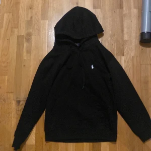 Svart hoodie från Polo Ralph Lauren - Svart hoodie från Polo Ralph Lauren med klassisk huva och snörning. Tröjan har lång ärm och en vit broderad logga på bröstet. Tillverkad i mjuk bomull för skön komfort. Perfekt för dig som gillar stilrena och enkla plagg med premiumkänsla.