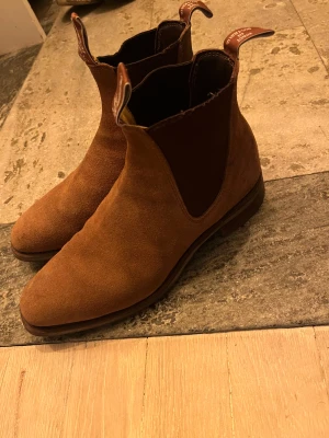 R.M. Williams - Säljer ett par äkta R.M. Williams boots, kända för sin tidlösa design, höga kvalitet och fantastiska komfort. Skorna är handgjorda i Australien i ett enda stycke läder, vilket ger både hållbarhet och en riktigt lyxig känsla. Bootsen är perfekta för både vardag och mer uppklätt, och de passar året runt. Nypris ligger vanligtvis runt 6 500 kr, så här har du chansen att fynda ett riktigt kvalitetspar!