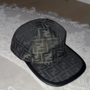 Svart keps från Fendi med FF-mönster - Snygg svart keps från Fendi med det klassiska FF-logomönstret över hela kepsen. Kepsen har böjd skärm och justerbar rem baktill med Fendi-logga. Tillverkad i bomullsmix och har en exklusiv känsla, perfekt för dig som vill sticka ut med designeraccessoar. (Pris kan diskuteras)