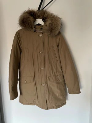 Beige parkas från Woolrich med pälskrage - Säljer en beige parkas från Woolrich med stor fluffig pälskant på huvan. Jackan har knappar framtill, två stora fickor med lock och två snedställda bröstfickor. Perfekt för kalla dagar och har en klassisk, stilren look.