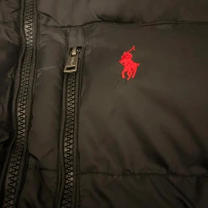 Svart pufferjacka från Polo Ralph Lauren - Svart pufferjacka från Polo Ralph Lauren med röd broderad logga på bröstet. Jackan har huva, dragkedja framtill och flera fickor med dragkedja. Materialet är glansigt och jackan är vadderad för extra värme. Perfekt för kalla dagar.