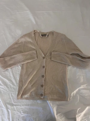 Beige ribbad kofta med knappar - Snygg beige kofta med ribbad struktur och v-ringning. Tröjan har långa ärmar och knäppning med fem matchande knappar framtill. Perfekt för lager-på-lager och enkel att styla med olika outfits. Mjuk och skön känsla, passar dig som gillar stilrena plagg.