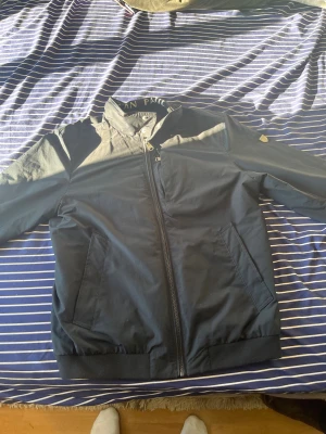 Svart bomberjacka från Jean Paul M - Snygg svart bomberjacka från Jean Paul med diskret logga på kragen och klassisk dragkedja framtill. Jackan har två snedställda fickor, ribbade muddar och en cool detalj med röd-vit-blått band på insidan. Perfekt för dig som gillar stilren och sportig look.