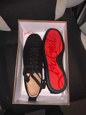 Christian Louboutin svarta sneakers - Säljer ett par svarta sneakers från Christian Louboutin i mocka med klassisk röd sula och snörning. Skorna har rund tå och diskreta metalldetaljer på sidan. Ikonisk design med lyxig känsla och snygg kontrast mellan svart ovandel och röd undersida. Priset kan diskuteras.