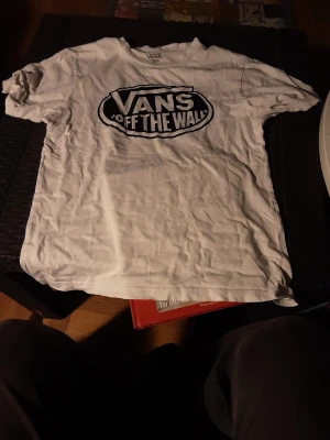 Vit Vans t-shirt med tryck - Säljer en vit t-shirt från Vans med det klassiska 'Off The Wall'-trycket i svart på bröstet. T-shirten har rund hals och korta ärmar, perfekt för dig som gillar streetwear och skatekänsla. Materialet är mjuk bomull och passformen är normal.