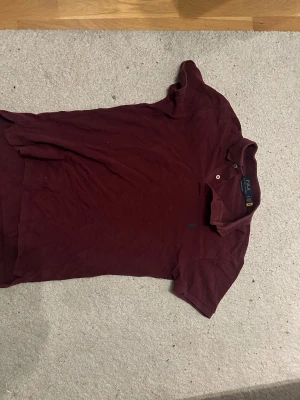 Polo Ralph Lauren piké - Säljer en vinröd pikétröja från Polo Ralph Lauren med klassisk krage och två knappar framtill. Tröjan har korta ärmar och den ikoniska broderade loggan på bröstet. Perfekt för dig som gillar stilrena och tidlösa plagg.