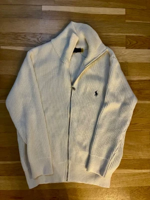 Vit stickad kofta från Polo Ralph Lauren - Säljer en ribbstickad vit kofta från Polo Ralph Lauren med dragkedja framtill och klassisk logga på bröstet. Tröjan har hög krage och långa ärmar, perfekt för att hålla värmen under kyliga dagar. Materialet är mjuk bomull och passformen är normal.