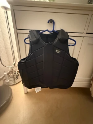 Svart säkerhetsväst från CRW - Svart säkerhetsväst från CRW med quiltad design och justerbara axelremmar. Västen har dragkedja och kardborre framtill för extra säkerhet och passform. Perfekt för ridning och ger bra skydd vid aktiviteter. Materialet är syntet och västen har en stilren, sportig look. PRIS KAN DISSKUTERAS