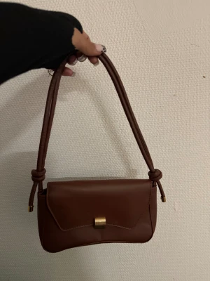 Brun axelväska med gulddetalj - Snygg brun axelväska i skinn med stilren design och guldfärgat lås framtill. Väskan har ett mjukt handtag med knutdetaljer och en enkel insida med plats för det viktigaste. Perfekt storlek för att ta med sig ut och matcha med olika outfits.