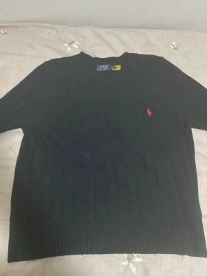 Svart kabelstickad tröja Polo Ralph Lauren - Svart kabelstickad tröja från Polo Ralph Lauren med klassisk rund hals och röd broderad logga på bröstet. Tröjan har ribbade muddar och är tillverkad i mjukt stickat material. Perfekt för dig som gillar stilren och tidlös design.