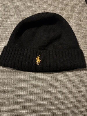 Svart mössa från Polo Ralph Lauren - Svart ribbstickad mössa från Polo Ralph Lauren med klassisk gul broderad logga framtill. Enkel och stilren design som passar perfekt till kalla dagar. Tillverkad i mjukt material för skön komfort och snygg passform. Man hittar inte den här sorten längre. Liknande produkter Kostar 695 kr på webbsidan och jag säljer för 300