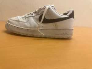 Nike Air Force 1 vita sneakers - Nike Air Force 1 sneakers i vitt skinn med svart swoosh på sidan. Klassisk låg modell med rund tå, snörning och perforeringar på tån för extra ventilation. Platt sula med Air-dämpning för skön komfort. Perfekt för dig som gillar streetwear och tidlös stil.