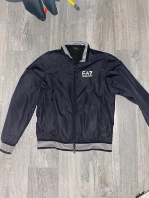Svart jacka från EA7 - Snygg svart jacka från EA7 Emporio Armani med vita ränder på muddar och krage. Jackan har dragkedja framtill, två fickor och logga på bröstet. Perfekt för dig som vill ha en stilren och sportig look. Är large men passar riktig bra på S&M (Det som står i ”storlek” är inte rätt storlek)
