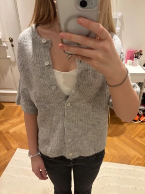 Grå kofta från Gina Tricot - Kortärmad grå kofta från gina tricot. Koftan har knappar framtill och är gjord i mjukt stickat material som känns skönt mot huden. Perfekt att slänga på över ett linne.