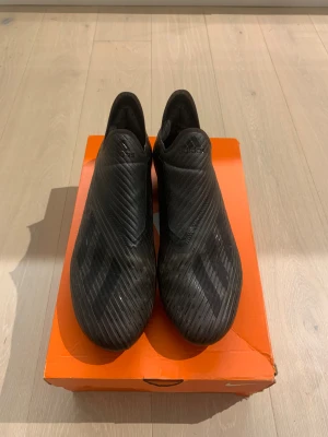 Svarta Adidas X fotbollsskor - Säljer ett par svarta Adidas X fotbollsskor med stilren design och diskreta ränder. Skorna har en strömlinjeformad form utan snörning, platt sula med dobbar och är tillverkade i syntetmaterial för lätt känsla på planen. Perfekt för dig som vill ha speed och kontroll. Storlek 42 2/3.