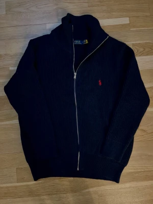 Svart stickad kofta från Polo Ralph Lauren - Svart ribbstickad kofta från Polo Ralph Lauren med röd broderad logga på bröstet. Tröjan har hel dragkedja framtill, hög krage och långa ärmar. Materialet är mjuk ull och passformen är normal. Perfekt för dig som gillar klassisk stil med en modern touch.
