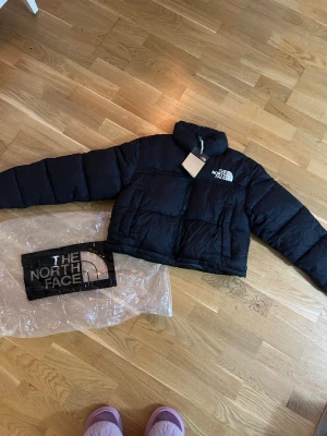 Svart pufferjacka från The North Face - Säljer en svart pufferjacka från The North Face med hög krage och dragkedja framtill. Jackan har en boxig passform och är vadderad för extra värme. Loggan är broderad på bröstet och jackan levereras med originalpåse.