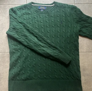 Grön stickad tröja Polo Ralph Lauren - Snygg grön stickad tröja från Polo Ralph Lauren med klassiskt kabelstickat mönster och broderad logga på bröstet. Rund hals och långa ärmar med ribbade muddar. Perfekt för dig som gillar stilrena och tidlösa plagg.