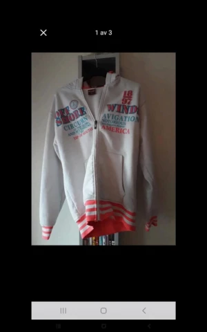 Vit hoodie med tryck och rödrandiga detaljer - Säljer en vit hoodie med färgglatt tryck framtill i blått och rött. Tröjan har dragkedja, huva och fickor. Muddar och nederkant har röda och vita ränder som ger en sportig vibe. Perfekt för dig som gillar streetwear och bekväma plagg.