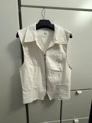 Vit jeansväst från Amisu med dragkedja - Snygg vit jeansväst från Amisu i storlek L. Västen har en klassisk krage och stängs med en silverfärgad dragkedja framtill. Materialet är stadigt och har en cool, lite boxig passform. Perfekt att styla över en t-shirt eller hoodie för en trendig look.