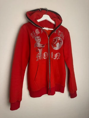 🔥Limited Röd hoodie med strasstenar Aim For The Moon - 🔥Limited Röd hoodie med dragkedja från Aim For The Moon. Framsidan har stora silvriga strasstenar i form av text och motiv, inklusive en djävul och en måne. Tröjan har huva, ribbade muddar och två fickor framtill. Perfekt för dig som vill ha något unikt och snyggt. Passar för både dam och herr. 📏Storlek:M ♻️Skick: Mycket bra, knappt använd (alla stenar sitter kvar). 📊Färg: Röd. ÖPPEN för bud!