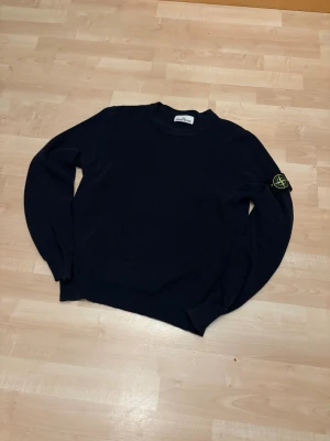 Mörkblå stickad tröja Stone Island - Säljer en mörkblå stickad tröja från Stone Island med klassisk rund hals och ribbade muddar. Tröjan har lång ärm och den ikoniska Stone Island-loggan på vänster ärm. Perfekt för dig som gillar stilrena och snygga plagg.