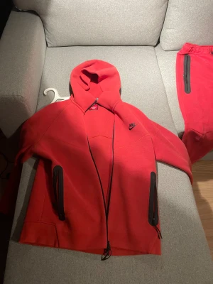 Röda Nike mjukisbyxor med dragkedja - Snygga röda mjukisbyxor från Nike med svarta detaljer och dragkedja på fickan. Byxorna har snörning i midjan och är tillverkade i mjukt bomullsmaterial. Perfekt för chill eller träning, med en modern look och bekväm passform. Har ett märke på byxorna fast i röd färg så märks inte mycket alls. 