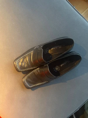 Svarta loafers i skinn från Boulevard - Snygga svarta loafers från Boulevard i äkta skinn med kontrasterande vita sömmar runt kanten. Modellen har en fyrkantig tå och platt sula, vilket ger en stilren och modern look. Perfekt för dig som gillar klassiska detaljer med en twist.