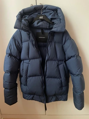 Mörkblå Peak Performance pufferjacka S - Säljer en mörkblå pufferjacka från Peak Performance med huva och quiltad design. Jackan har lång ärm, dragkedja framtill och en stilren look som passar perfekt när det är kallt ute. Materialet är syntet och dun, vilket gör den både varm och lätt.