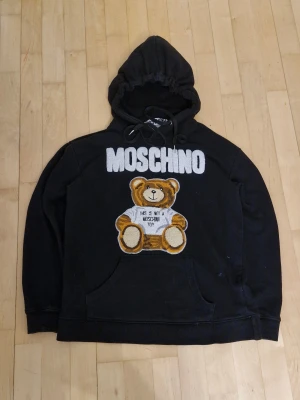 Svart Moschino hoodie med nalle - Svart hoodie från Moschino med stor broderad nalle på framsidan och vit Moschino-text. Tröjan har huva med snörning och känguruficka. Materialet är 100% bomull med detaljer i viskos och silke. Perfekt för dig som gillar statement-plagg och coola designer.