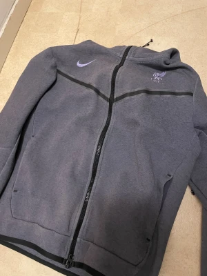 Grå Nike Liverpool hoodie med dragkedja - Säljer en grå hoodie från Nike med Liverpool FC-logga och lila swoosh på bröstet. Jackan har hel dragkedja, huva med snörning och svarta detaljer längs bröstet. Mjuk och skön i bomullsmix, perfekt för dig som gillar sportig stil.