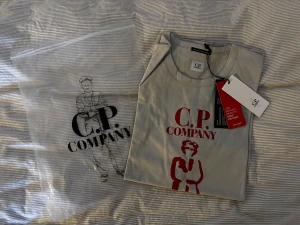 Grå C.P. Company t-shirt med tryck - Snygg grå t-shirt från C.P. Company med rött tryck framtill och klassisk rund hals. T-shirten är i mjuk bomull och har en stilren look med logga och grafiskt motiv. Perfekt för dig som gillar streetwear och vill ha något från ett ikoniskt märke.
