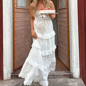 Vit långklänning med volanger - Superfin vit långklänning i bomull med broderade hålmönster, volanger och omlottkänsla. Klänningen har smala knytband i nacken, öppen rygg och v-ringad front. Endast använd en gång! buda vid intresse❣️