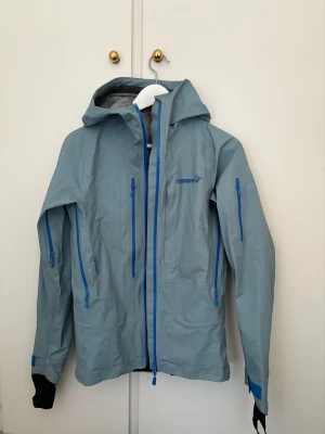 Blå vindjacka från Norrøna - Snygg ljusblå skidjacka från Norrøna med huva och flera blå dragkedjor. Jackan har lång ärm, hög krage och praktiska fickor på bröstet och ärmarna. Perfekt för utomhusaktiviteter och äventyr. Modellen är stilren och funktionell. Modell - Lofoten. 