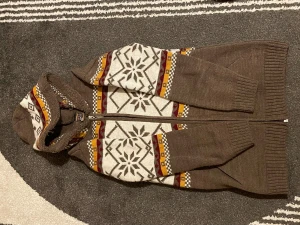 Brun hoodie med vintermönster och huva - Säljer en brun hoodie från Alpaca of Sweden med vit, röd och gul snöflingemönster. Tröjan har huva, dragkedja och ribbade muddar. Stickad i mjukt material, perfekt för kalla dagar. Snygga detaljer och skön passform.