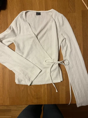 Vit omlottopp från Gina Tricot - Säljer en vit omlottopp från Gina Tricot i storlek S. Toppen har långa ärmar, knyt i sidan och ett ribbat, strukturerat tyg som ger en snygg känsla. Perfekt för dig som gillar stilrena och enkla plagg med lite extra detalj.