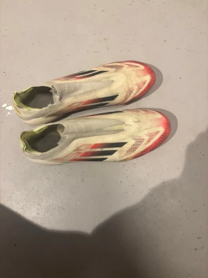 Adidas f50 fotbollsskor - Säljer ett par Adidas f50 fotbollsskor i vitt med röda och svarta detaljer samt inslag av gult. Skorna har en strömlinjeformad design utan snörning, platt sula med dobbar och är tillverkade i syntetmaterial för lätthet och snabbhet på planen.