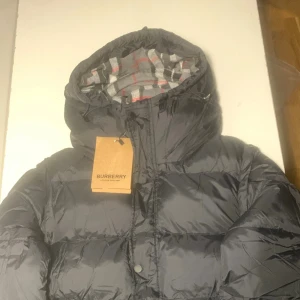 Burberry jacka - Snygg svart dunjacka från Burberry med stor huva och klassiskt rutigt foder i grått, vitt och rött. Skriv gärna om du vill diskutera om priset!