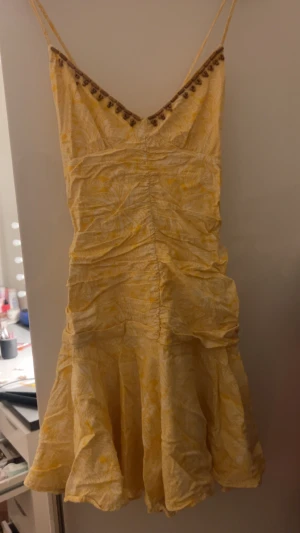 Gul mönstrad kortklänning från Zara - Superfin gul kortklänning från Zara med vitt paisleymönster och tunna axelband. Klänningen har snygga träpärlor längs v-ringningen och rynkade detaljer framtill. Materialet är mjukt bomullstyg, perfekt för varma dagar. Fladdrig kjol och omlottkänsla upptill.