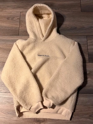 Beige fleece hoodie Jack & Jones - Mysig beige hoodie från Jack & Jones Originals Studio med fluffig fleeceyta och broderad logga på bröstet. Tröjan har huva, ribbade muddar och långärmad design. Perfekt för kalla dagar när du vill vara både chill och varm.