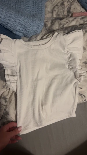 Vit volangtopp från Zara - Supersöt vit topp från Zara med volangdetaljer på axlarna. Ribbad struktur och rund halsringning. Perfekt för dig som vill ha en enkel men ändå extra topp till din outfit.