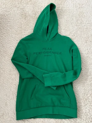 Grön hoodie från Peak Performance - Snygg grön hoodie från Peak Performance med broderad logga på bröstet. Tröjan har huva, magficka och långa ärmar med ribbade muddar. Perfekt för chill eller när du vill ha en sportig look.