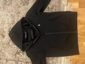 Svart hoodie från Polo Ralph Lauren S/M - Svart hoodie från Polo Ralph Lauren med dragkedja och huva. Klassisk design med vit broderad logga på bröstet och två fickor framtill. Mjuk insida och normal passform, perfekt för chill dagar. Materialet är bomull och polyester.