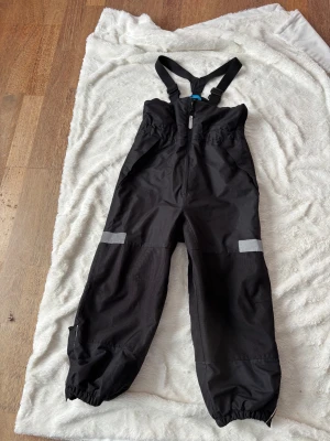 Svarta täckbyxor med hängslen 116 - Svarta täckbyxor från Åhléns med justerbara hängslen, dragkedja framtill och reflexdetaljer på benen. Byxorna har resår vid bensluten och är gjorda i slitstarkt syntetmaterial, perfekta för kalla dagar utomhus. Spår av användning syns på hö knä och på rumpan , syns i bilderna . Inget trasigt, tom kardborreband i bra skick