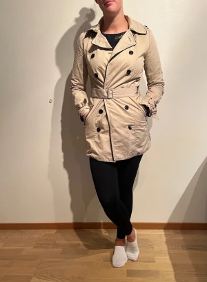 Beige trenchcoat med gulddetaljer - Skick: Fint. En trasig knapp och ej original-skärp. 2 nya knappar medföljer   Tvättråd: 30° 