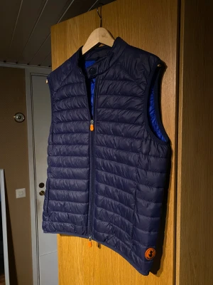 Mörkblå dunväst från Save The Duck - Snygg mörkblå dunväst från Save The Duck med horisontella quiltade sömmar med en hög och unik krage. Västen har dragkedja framtill med orange detalj och en rund orange logotyp på sidan. Perfekt för lager-på-lager och stilren look.