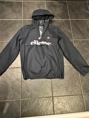Mörkblå vindjacka från Ellesse med huva - Marinblå vindjacka från Ellesse med stor logga framtill, huva och halv dragkedja. Jackan har meshfoder på insidan och är perfekt för blåsiga dagar. Snygg och sportig stil med klassisk Ellesse-detalj på dragkedjan. Storlek 158-164/ 13-14. Ungefär storlek XS
