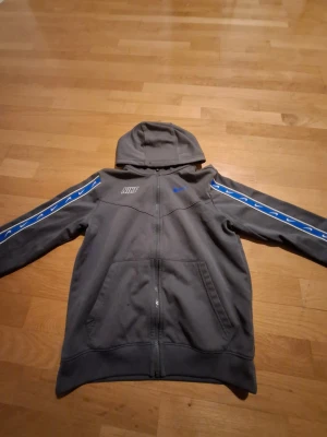Grå Nike hoodie med blå detaljer - Nike Dri-Fit hoodie i grått med blå och vita detaljer längs ärmarna. Tröjan har huva, dragkedja framtill och två fickor. Snyggt Nike-tryck på bröstet och ärmarna. Perfekt för träning eller chill. Materialet är polyester.