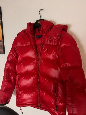 Röd dunjacka från Polo Ralph Lauren - Riktigt fet röd dunjacka från Polo Ralph Lauren med huva och dragkedjor på fickorna. Klassisk pufferstil med broderad logga på bröstet och hög krage. Perfekt för kalla dagar när du vill sticka ut och hålla värmen.