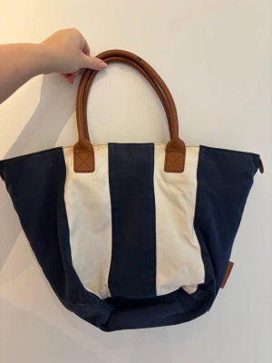 Randig shoppingväska med bruna handtag - Stor och rymlig shoppingväska med breda marinblå och vita ränder. Väskan har två stadiga handtag i brunt skinn som ger en snygg kontrast mot det randiga tyget. Perfekt för att bära med sig allt man behöver under dagen.
